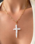 A close-up of a person wearing a 14k gold filled chain necklace with a textured white cross pendant, styled with a cream ribbed cardigan, by Camille Jewelry