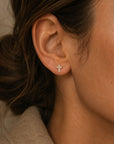 Close-up of an ear wearing a diamond cross stud earrings in 14K yellow gold with a neutral background by Camille Jewelry.