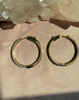 A pair of gold pave hinged hoop earrings, resting on a textured beige fabric surface with a crystal cluster in the background by Camille Jewelry