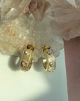 A pair of gold ribbed pave heart gem huggie hoop earrings, displayed on a white surface next to a pink fabric strip and clear quartz crystals by Camille Jewelry.