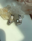A pair of sterling silver ribbed hoop huggie earrings with heart shaped cubic zirconia stone, displayed on a white surface with crystal clusters in the background by Camille Jewelry.