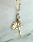 A gold necklace with a paperclip chain and three charms: a cross, a small clear rectangular stone, and a coffin-shaped pendant with 'L ♥ A' engraved and a border of small clear stones, displayed on a light marble surface.