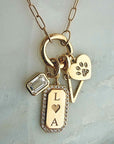 A gold-tone necklace with a paperclip chain and multiple charms, including a rectangular clear stone charm, a rectangular plate engraved with 'L A' and a heart, a triangular charm with a paw print, and a heart-shaped charm with a paw print, displayed on a light gray marble surface.