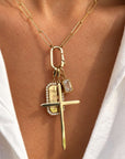A close-up of a 14K gold necklace worn on a person's neck, featuring a paperclip chain, a rectangular ring clasp, and a multi-pendant charm set with a cross, a emerald cut engraved pendant, and a clear diamond charm.