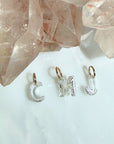 Three lab created floating diamond letter initial charms spelling 'CMJ' with 14k yellow gold jump rings, displayed on a light surface with rose quartz crystals in the background, by camille Jewelry