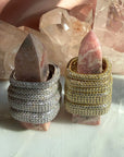 Two square ring styles displayed on pink stone, set against a background of clear quartz crystals. One ring is rhodium plated silver with pavé-set clear stones, and the other is gold plated silver with pavé-set stones by Camille Jewelry