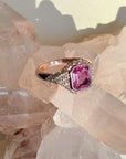 A 14k rose gold ring with a large emerald-cut pink gemstone and pavé-set clear stones, displayed on a cluster of rose quartz crystals by Camille Jewelry
