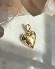 A polished 14k yellow gold puffy heart charm with natural diamonds, displayed on a light marble surface with crystal clusters in the background.