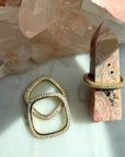 Pave gold plated sterling silver square rings, displayed on a light marble surface with a pink stone agate tower in the background, Camille Jewelry.