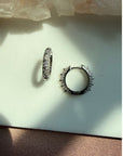 A pair of sterling silver studded pave hoop earrings, displayed on a light surface with natural shadows by Camille Jewelry.