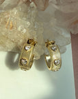 A pair of gold ribbed pave heart gem huggie hoop earrings, displayed on a white surface next to a pink fabric strip and clear quartz crystals by Camille Jewelry.