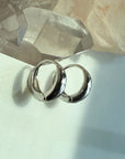 Two polished silver hoop earrings resting on a light surface with a translucent crystal cluster in the background by Camille Jewelry