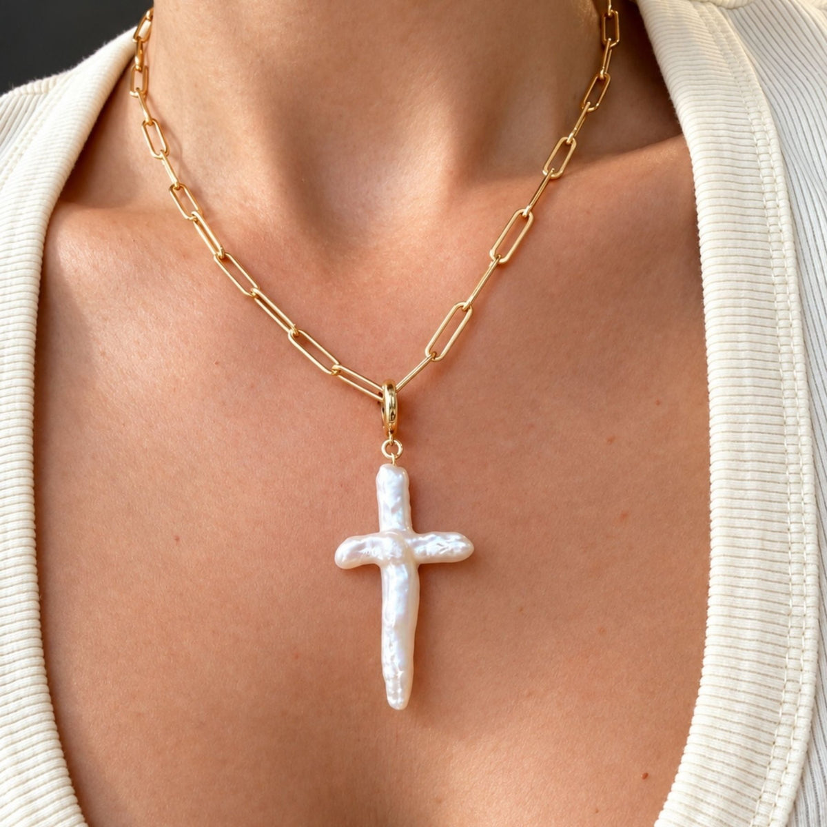 A close-up of a person wearing a 14k gold filled chain necklace with a textured white cross pendant, styled with a cream ribbed cardigan, by Camille Jewelry