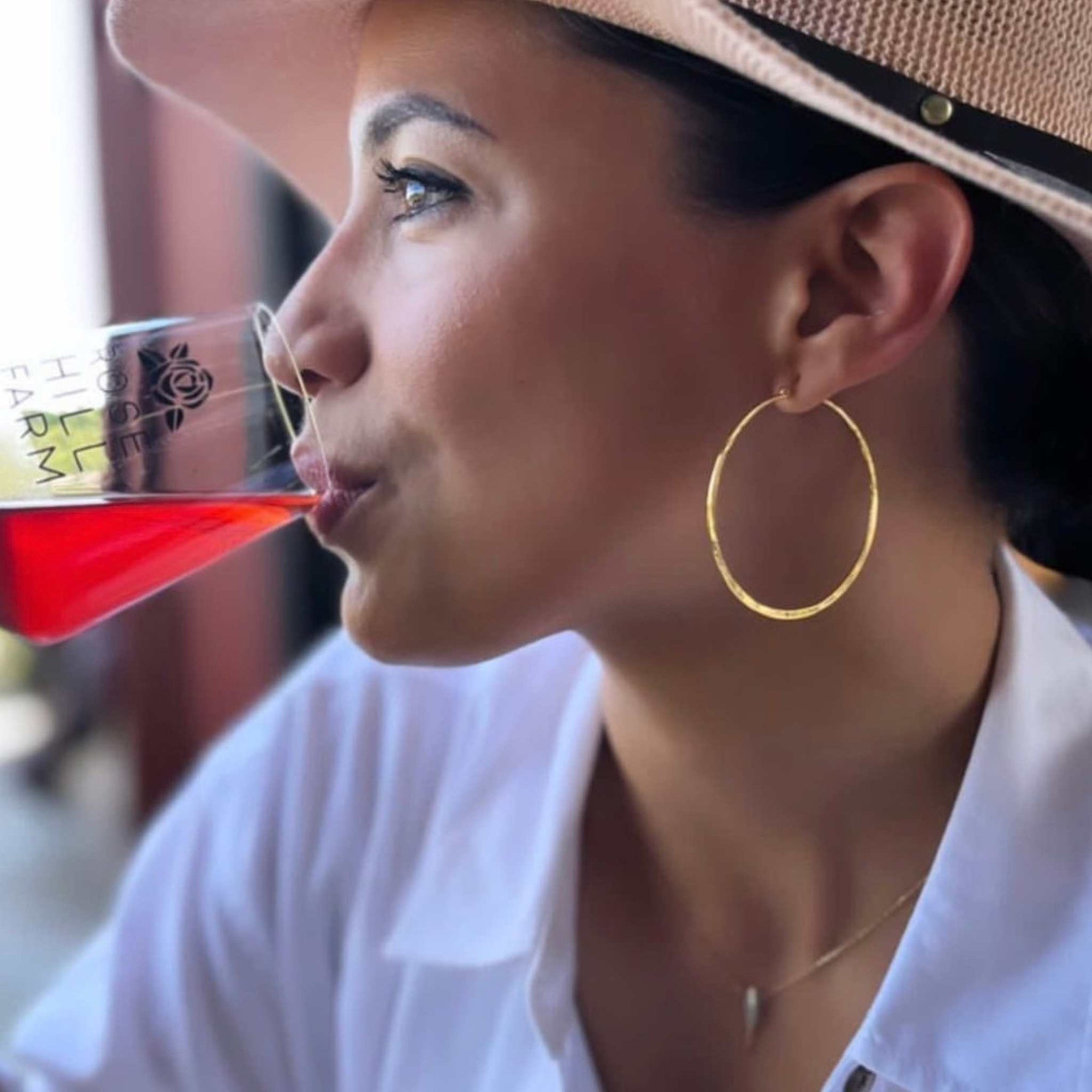 Gold hoop earrings on model sipping wine wearing a hat - Camille Jewelry