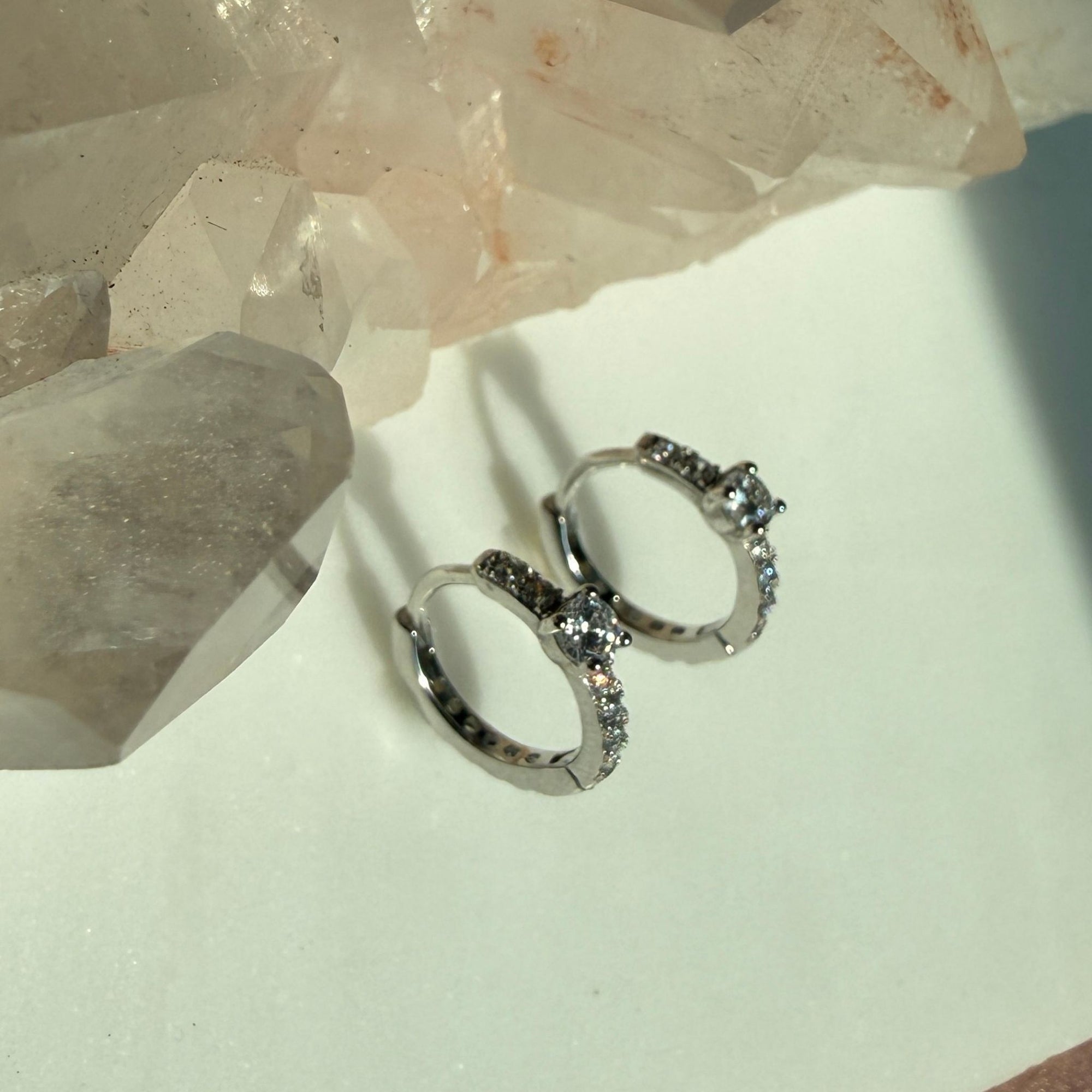 A pair of silver center stone pave hoop earrings with small clear stones, displayed on a light surface next to a large crystal cluster by Camille Jewelry.