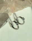 A pair of silver center stone pave hoop earrings with small clear stones, displayed on a light surface next to a large crystal cluster by Camille Jewelry.