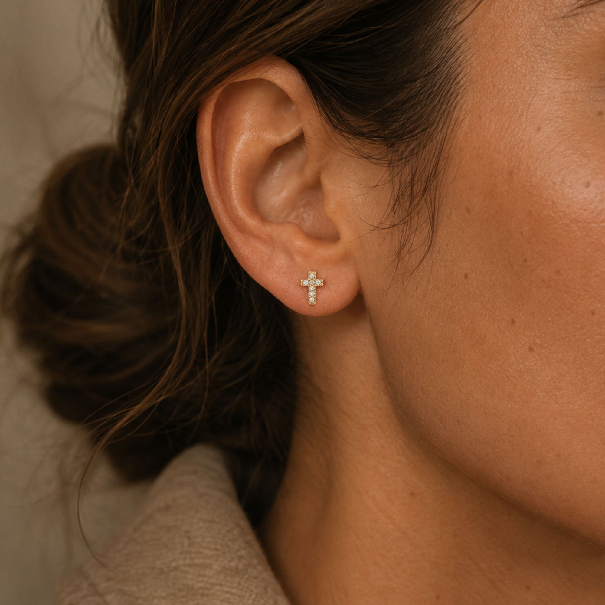 Close-up of an ear wearing a diamond cross stud earrings in 14K yellow gold with a neutral background by Camille Jewelry. 