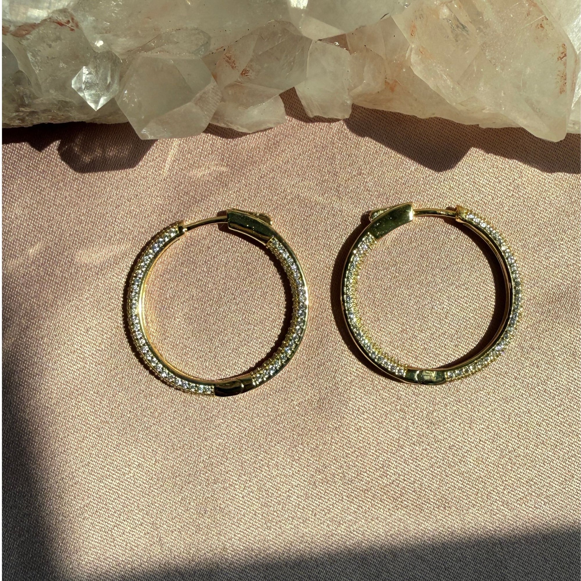 A pair of gold pave hinged hoop earrings, resting on a textured beige fabric surface with a crystal cluster in the background by Camille Jewelry