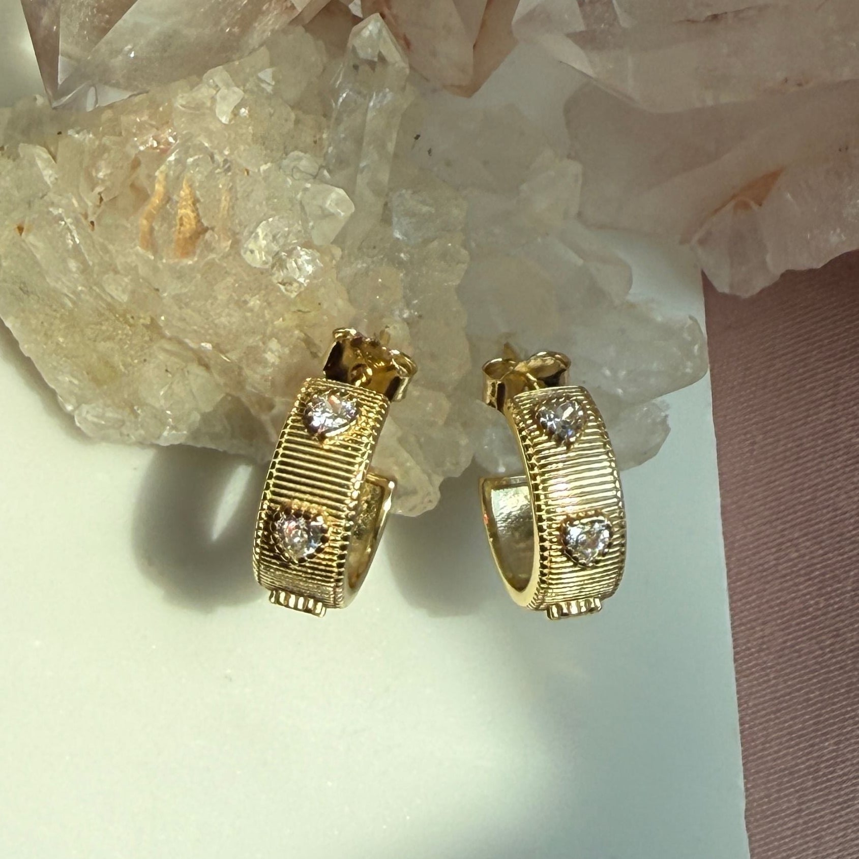 A pair of gold ribbed pave heart gem huggie hoop earrings, displayed on a white surface next to a pink fabric strip and clear quartz crystals by Camille Jewelry. 