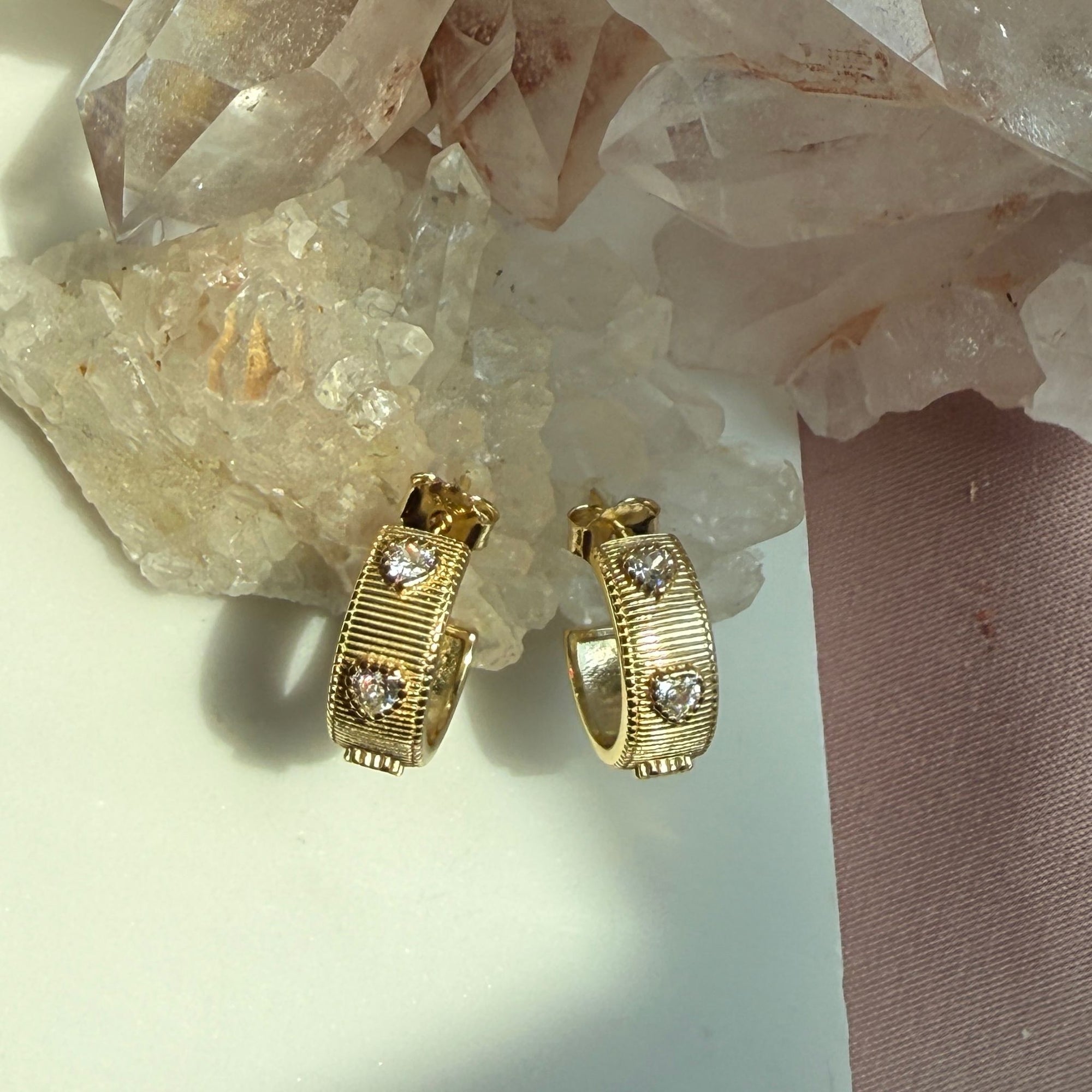 A pair of gold ribbed pave heart gem huggie hoop earrings, displayed on a white surface next to a pink fabric strip and clear quartz crystals by Camille Jewelry. 