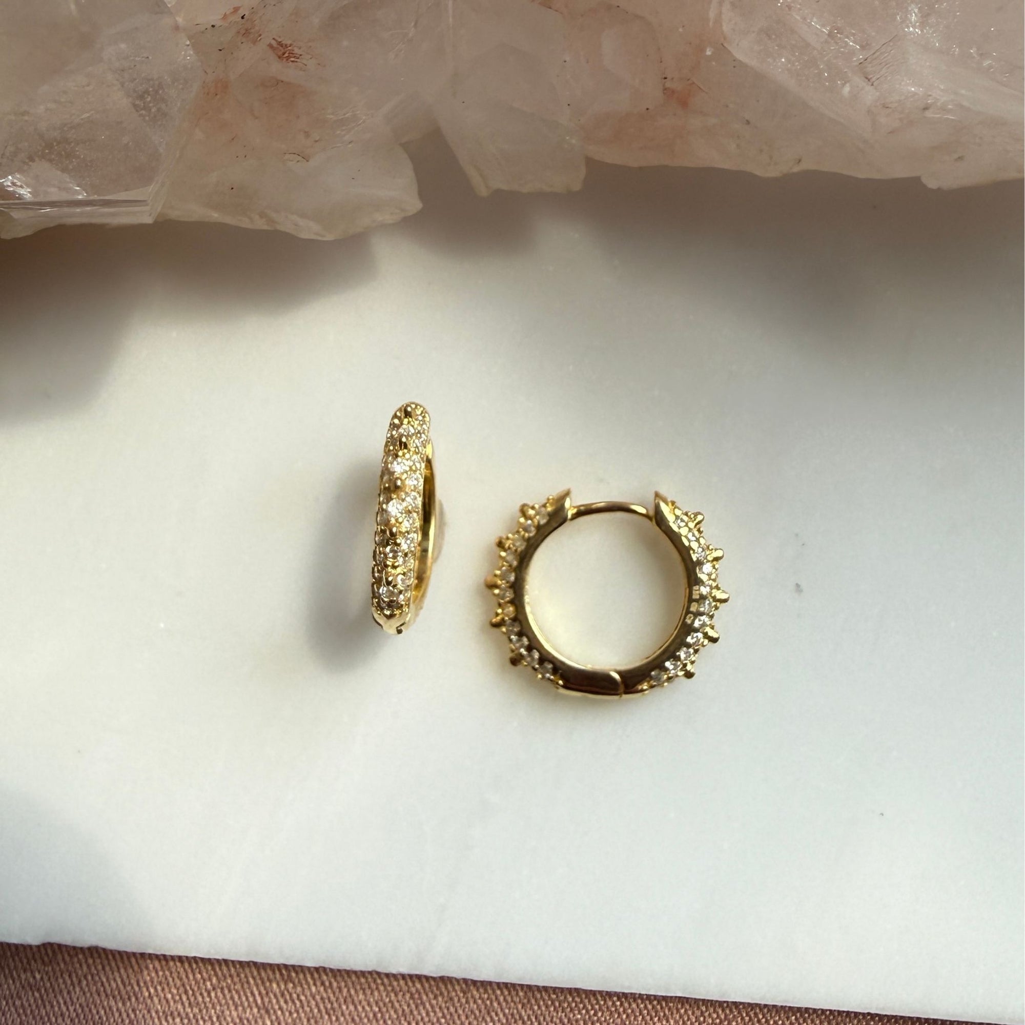 A pair of small gold studded pave hoop earrings with textured detailing, displayed on a white surface with a pink crystal cluster in the background by Camille Jewelry.