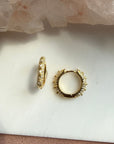 A pair of small gold studded pave hoop earrings with textured detailing, displayed on a white surface with a pink crystal cluster in the background by Camille Jewelry.
