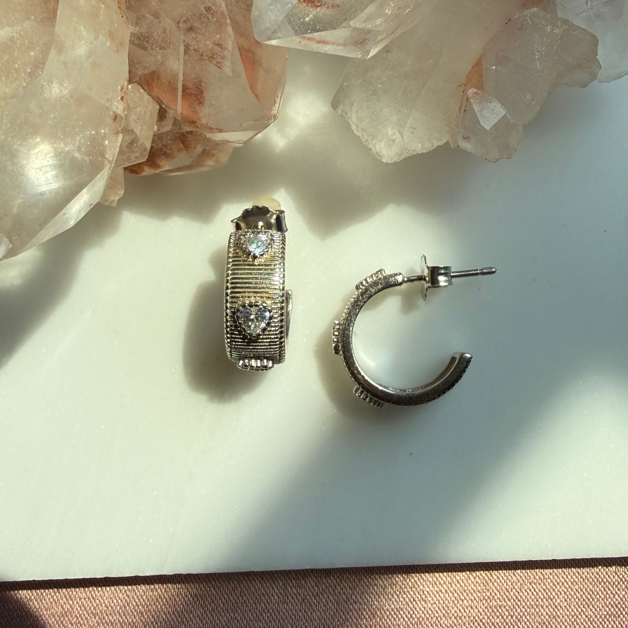 A pair of sterling silver textured ribbed hoop earrings with heart gem, displayed on a white surface with crystal clusters in the background by Camille Jewelry. 