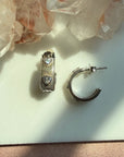 A pair of sterling silver textured ribbed hoop earrings with heart gem, displayed on a white surface with crystal clusters in the background by Camille Jewelry. 