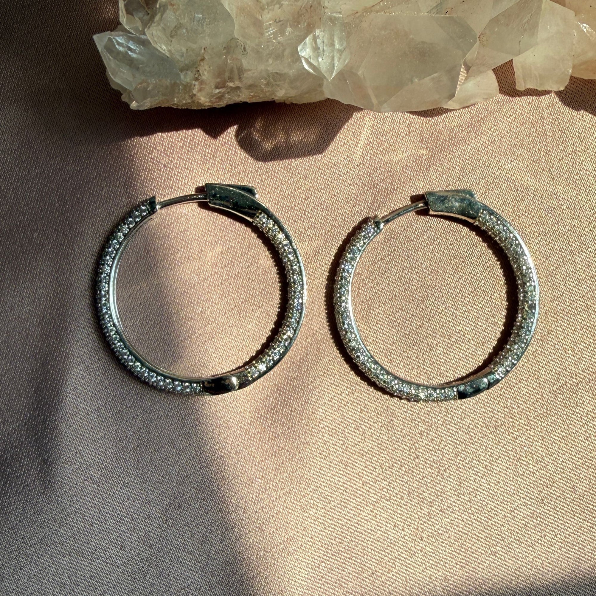 A pair of .925 sterling silver pave hinged hoop earrings, resting on a textured beige fabric surface with a crystal cluster in the background by Camille Jewelry
