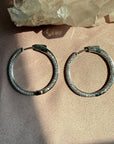 A pair of .925 sterling silver pave hinged hoop earrings, resting on a textured beige fabric surface with a crystal cluster in the background by Camille Jewelry