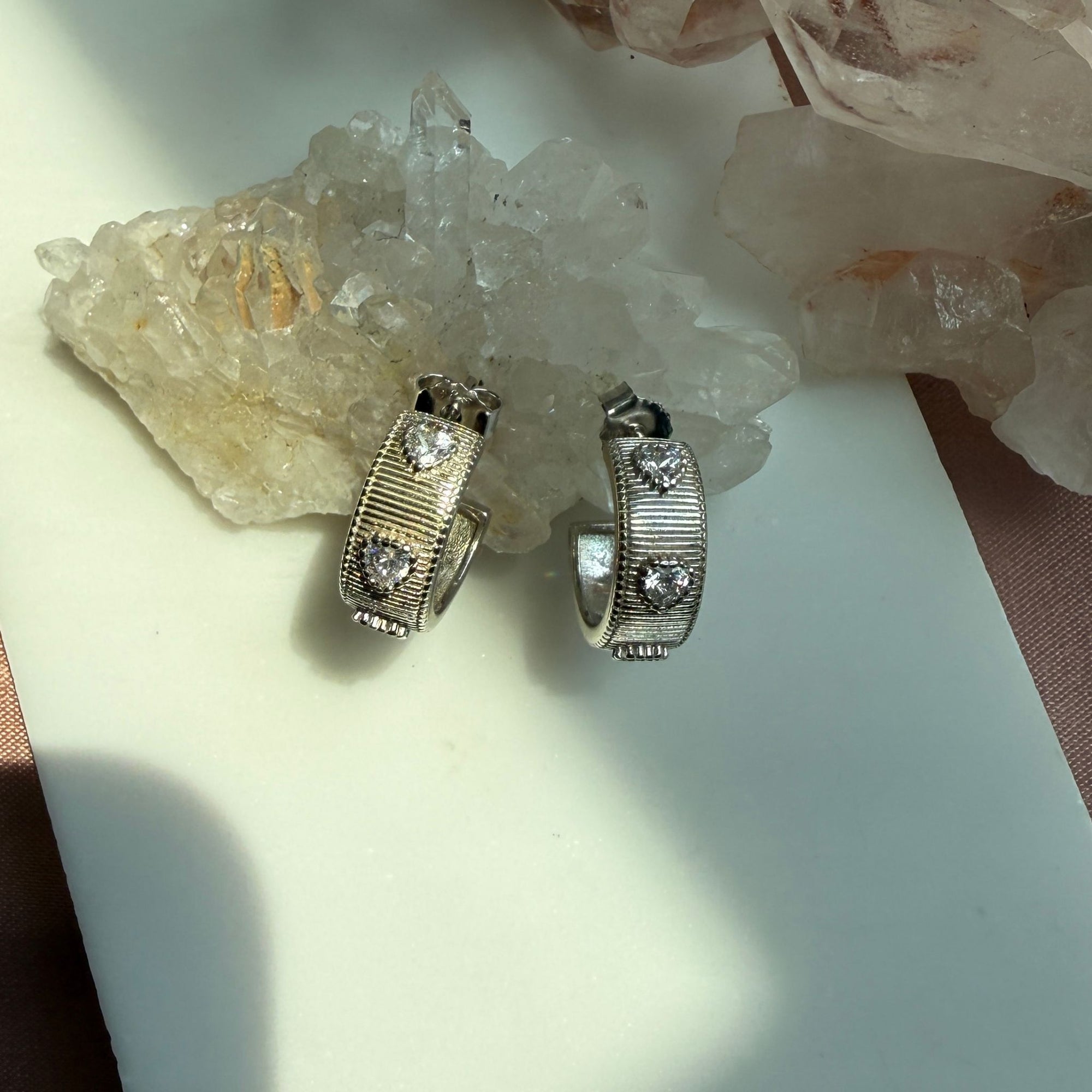 A pair of sterling silver ribbed hoop huggie earrings with heart shaped cubic zirconia stone, displayed on a white surface with crystal clusters in the background by Camille Jewelry. 