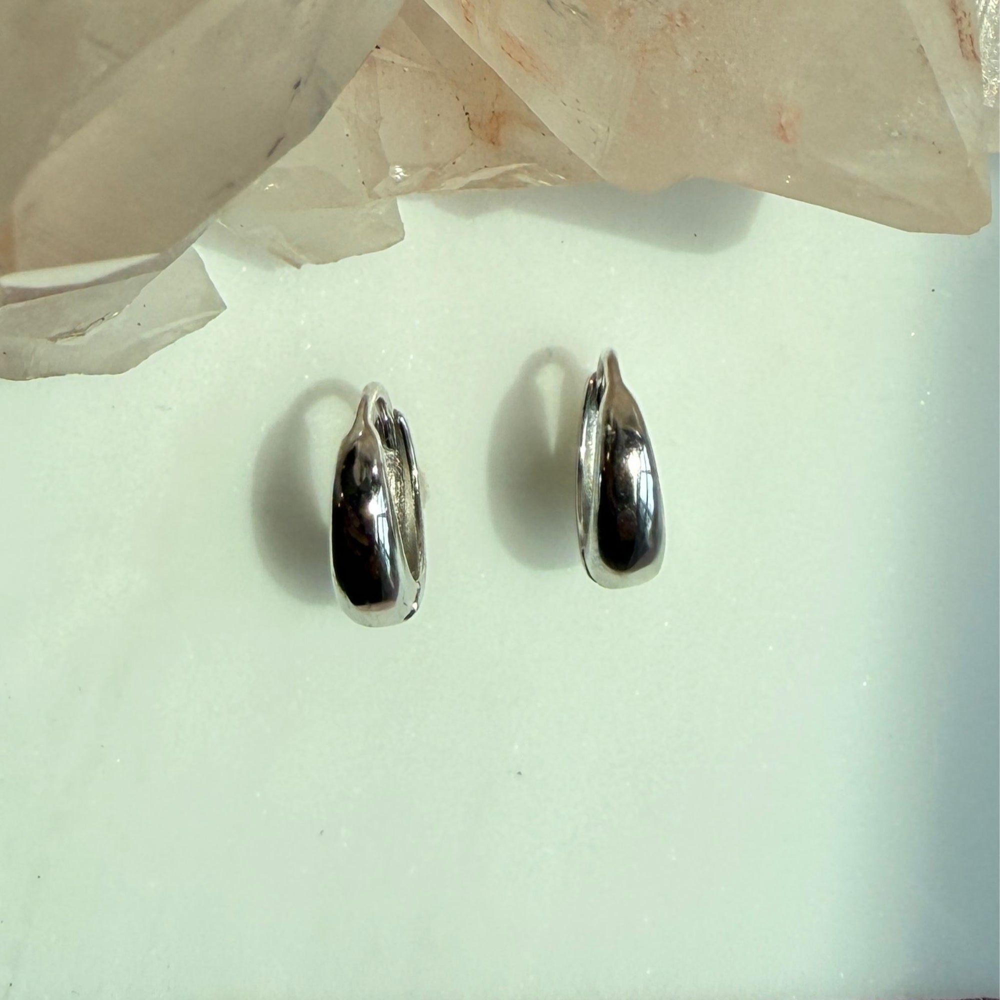 A pair of polished hinged silver huggie earrings, displayed on a light mint green surface with a translucent crystal cluster in the background.