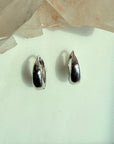A pair of polished hinged silver huggie earrings, displayed on a light mint green surface with a translucent crystal cluster in the background.