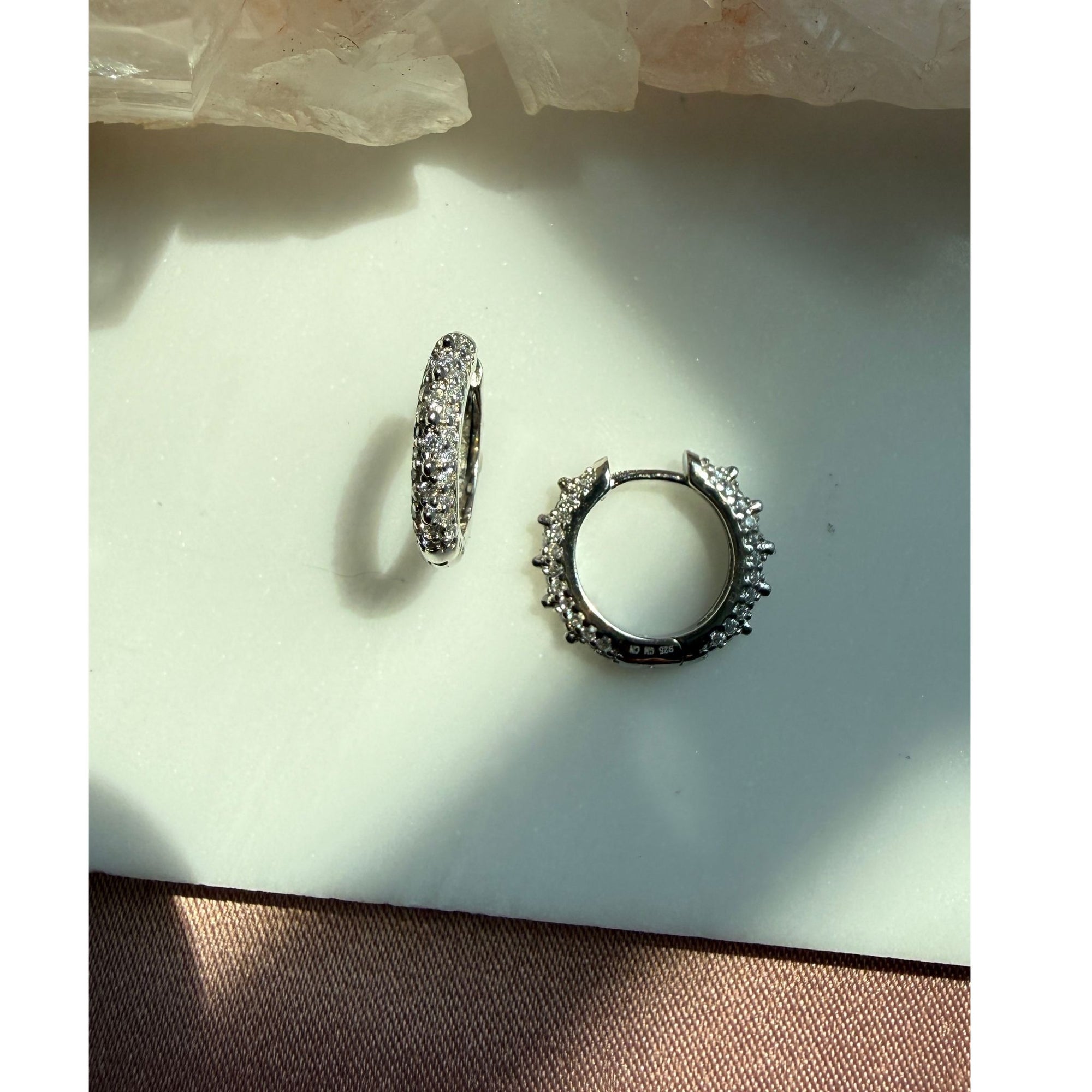 A pair of sterling silver studded pave hoop earrings, displayed on a light surface with natural shadows by Camille Jewelry.