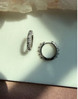 A pair of sterling silver studded pave hoop earrings, displayed on a light surface with natural shadows by Camille Jewelry.