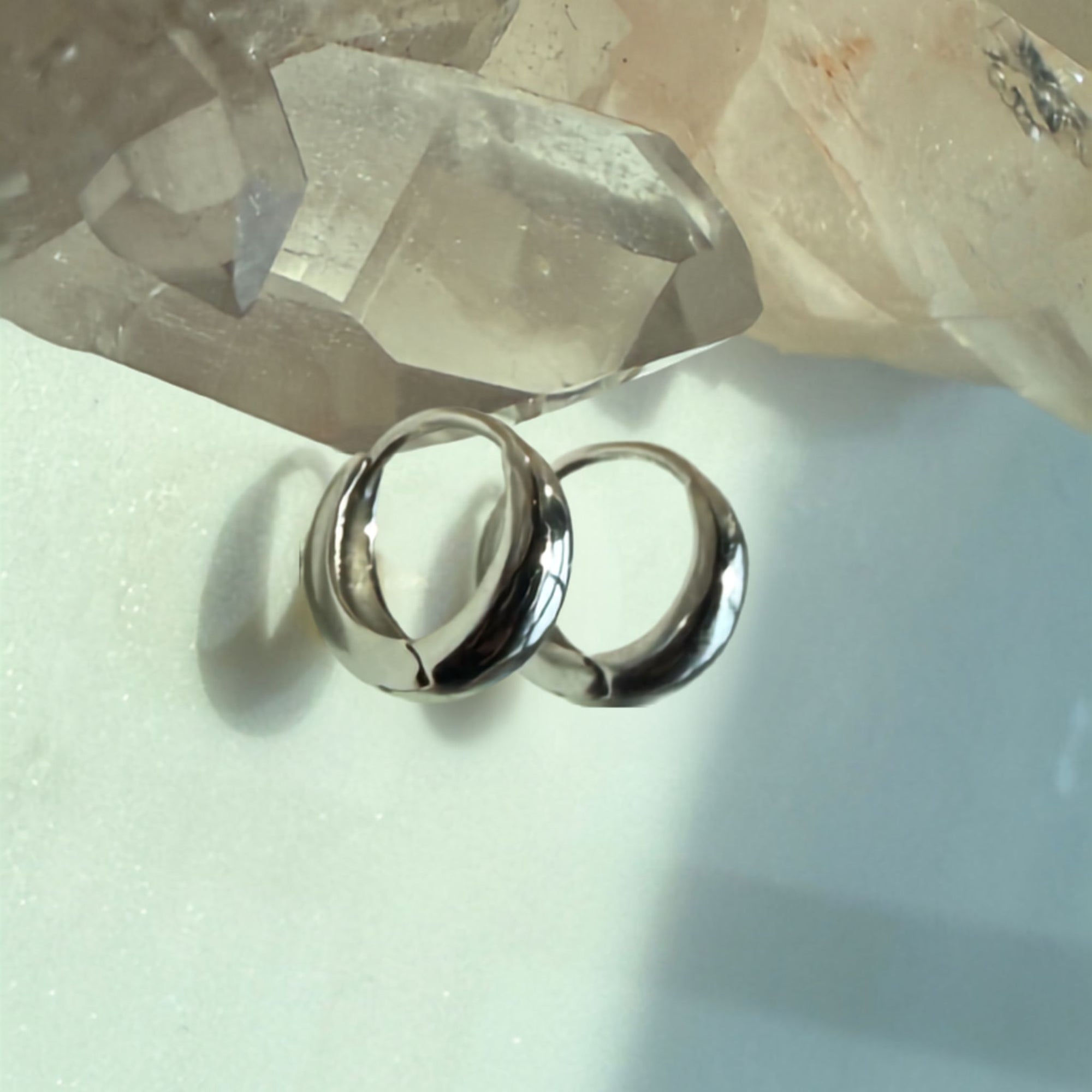 Two polished silver hoop earrings resting on a light surface with a translucent crystal cluster in the background by Camille Jewelry 