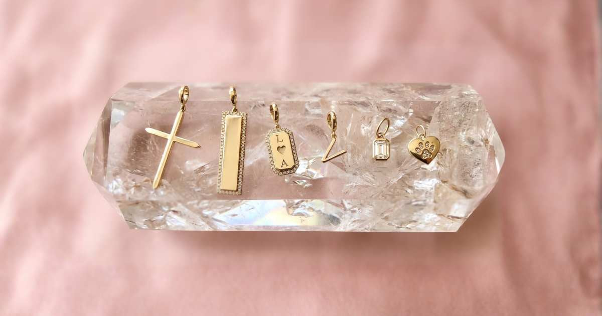 Five small 14k yellow gold jewelry charms displayed on a clear faceted crystal point, set against a soft pink fabric background for Camille Jewelry.