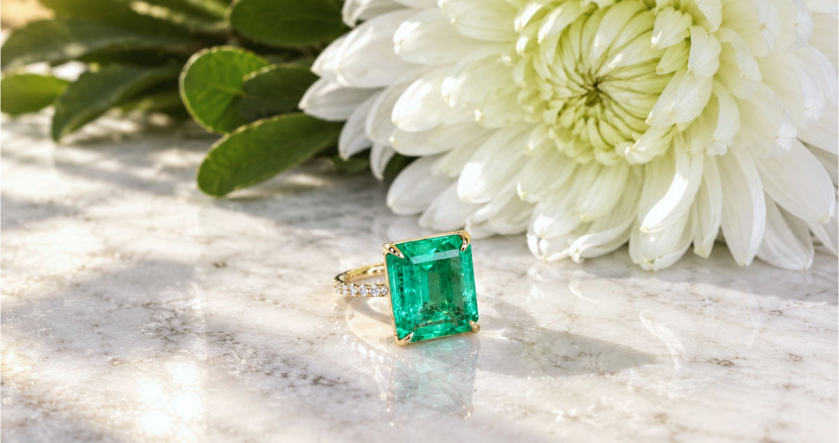 A 18K yellow gold ring featuring a large emerald-cut green gemstone with natural diamond band ring, styled on a white marble surface next to a white flower and green leaves, by Camille Jewelry