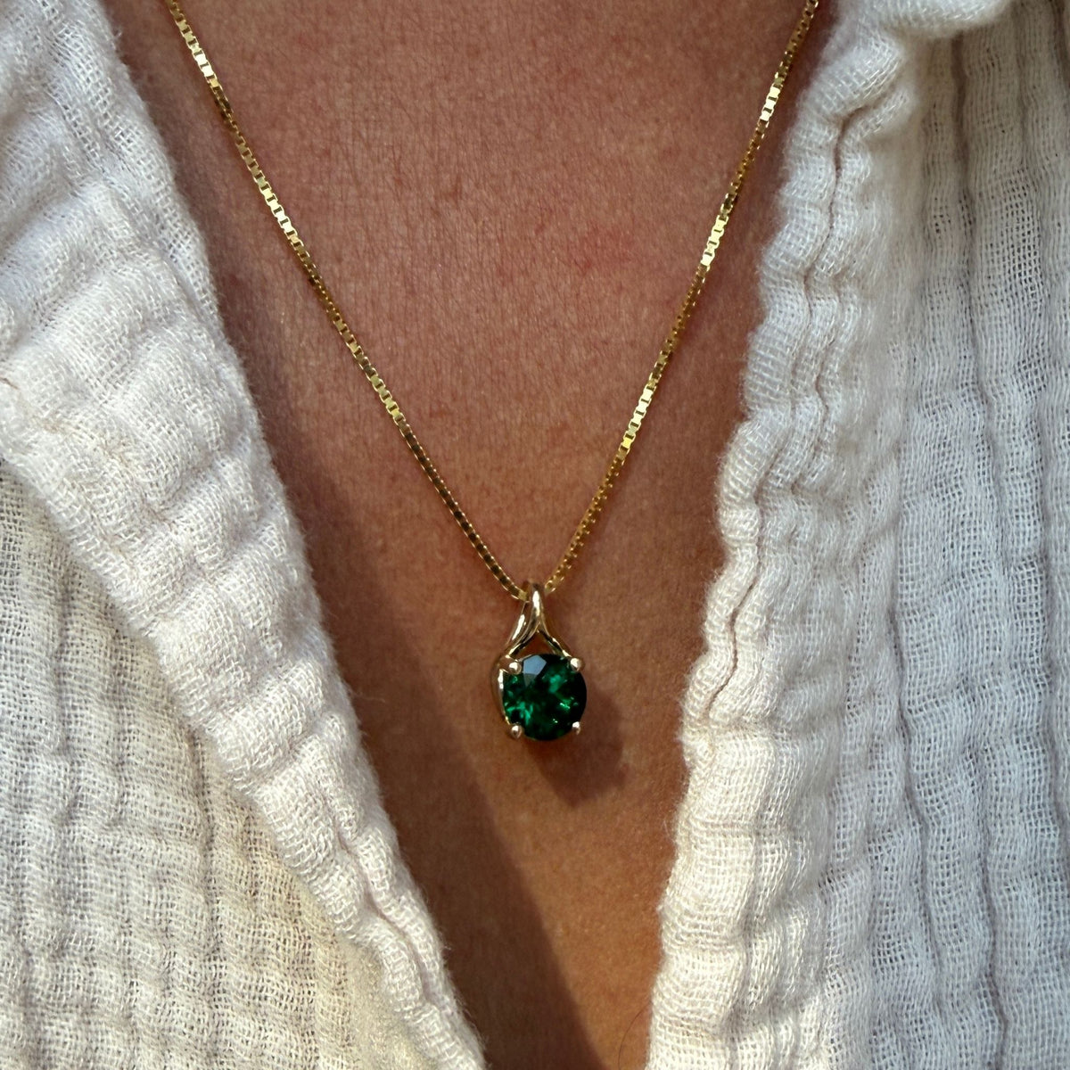 A close-up of a gold chain necklace with a teardrop-shaped green emerald pendant, worn on a person's neck over a white textured knit top by Camille Jewelry