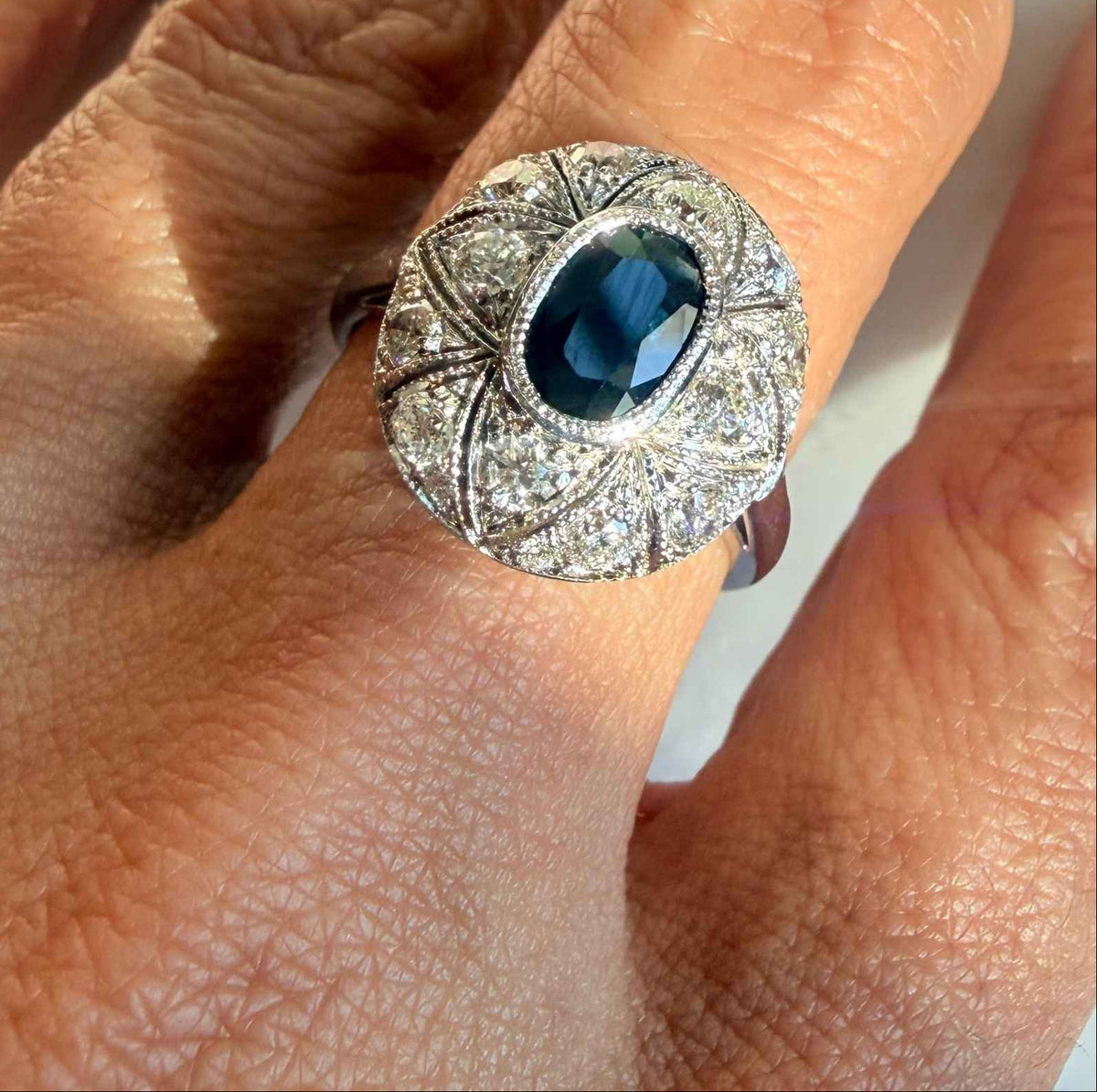 A close-up of a hand wearing a vintage-style round ring with sapphire center stone and a surrounding 14K white gold milgrain setting by Camille Jewelry.