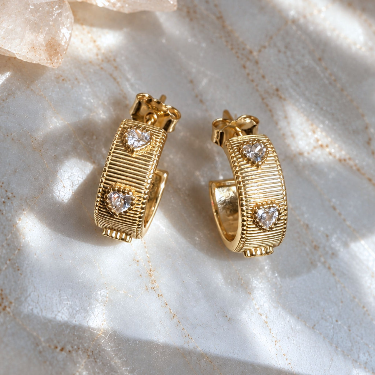 A pair of gold huggie hoop earrings with textured ribbed bands and three clear heart-shaped cubic zirconia on each earring, displayed on a light marble surface with soft shadows, by Camille Jewelry