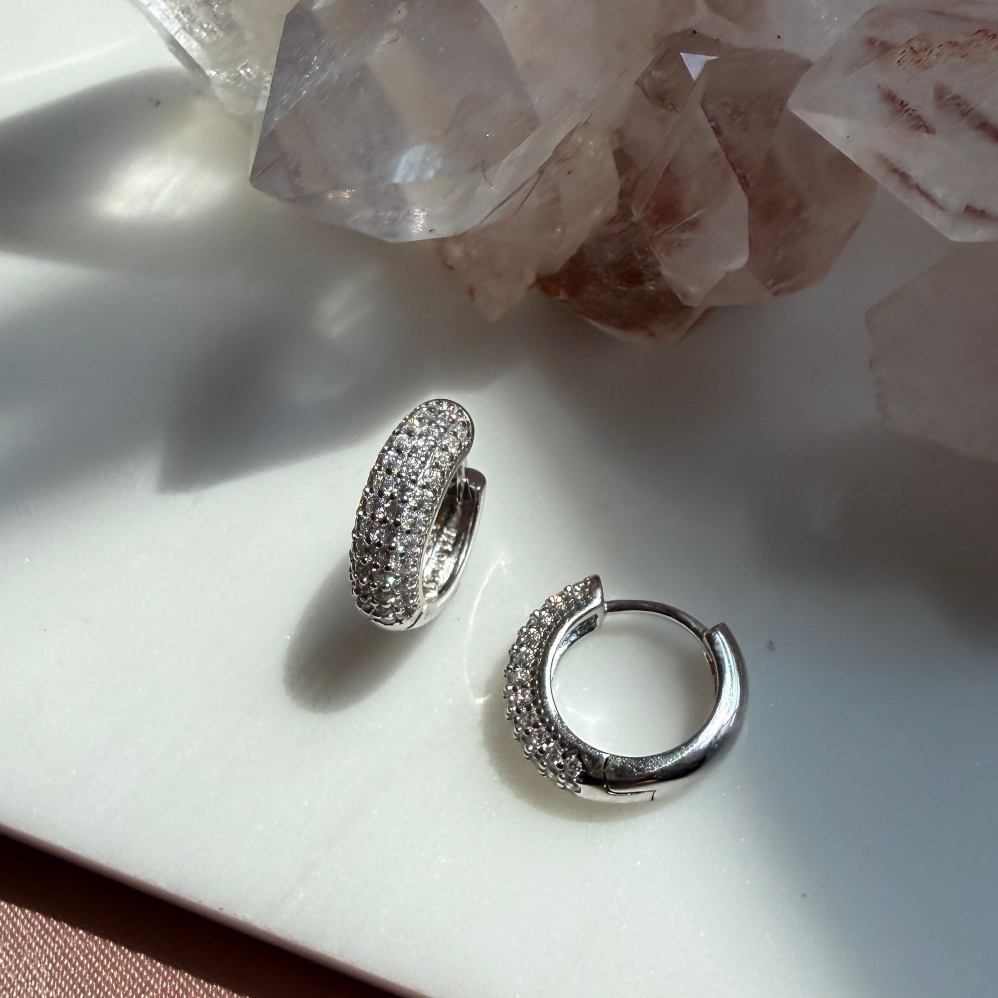 A pair of silver domed pave hoop earrings covered in clear pavé stones, displayed on a white surface with pink crystal clusters in the background by Camille Jewelry
