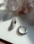 A pair of silver domed pave hoop earrings covered in clear pavé stones, displayed on a white surface with pink crystal clusters in the background by Camille Jewelry