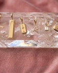 Six gold jewelry for women charms in solid 14k gold and diamonds displayed on a clear faceted crystal holder, set against a soft dusty pink suede background by Camille Jewelry. 