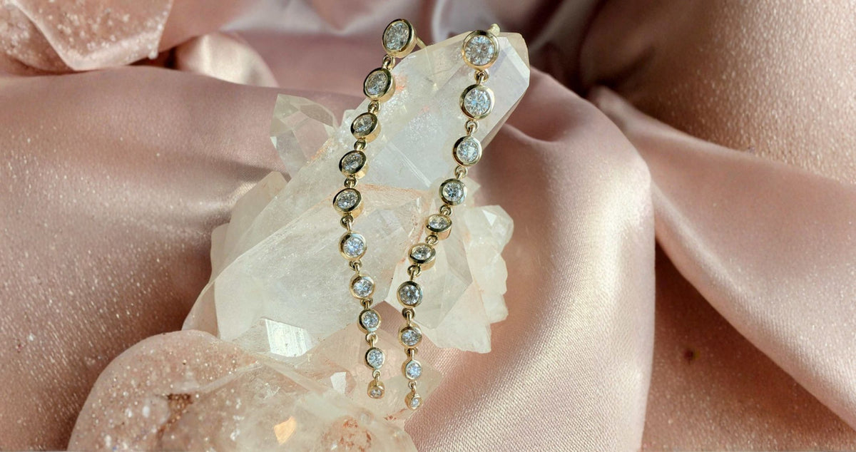A pair of 14K yellow gold earrings with lab diamonds that are bezel set and graduate, displayed on a white crystal cluster over soft blush pink satin fabric by Camille Jewelry.