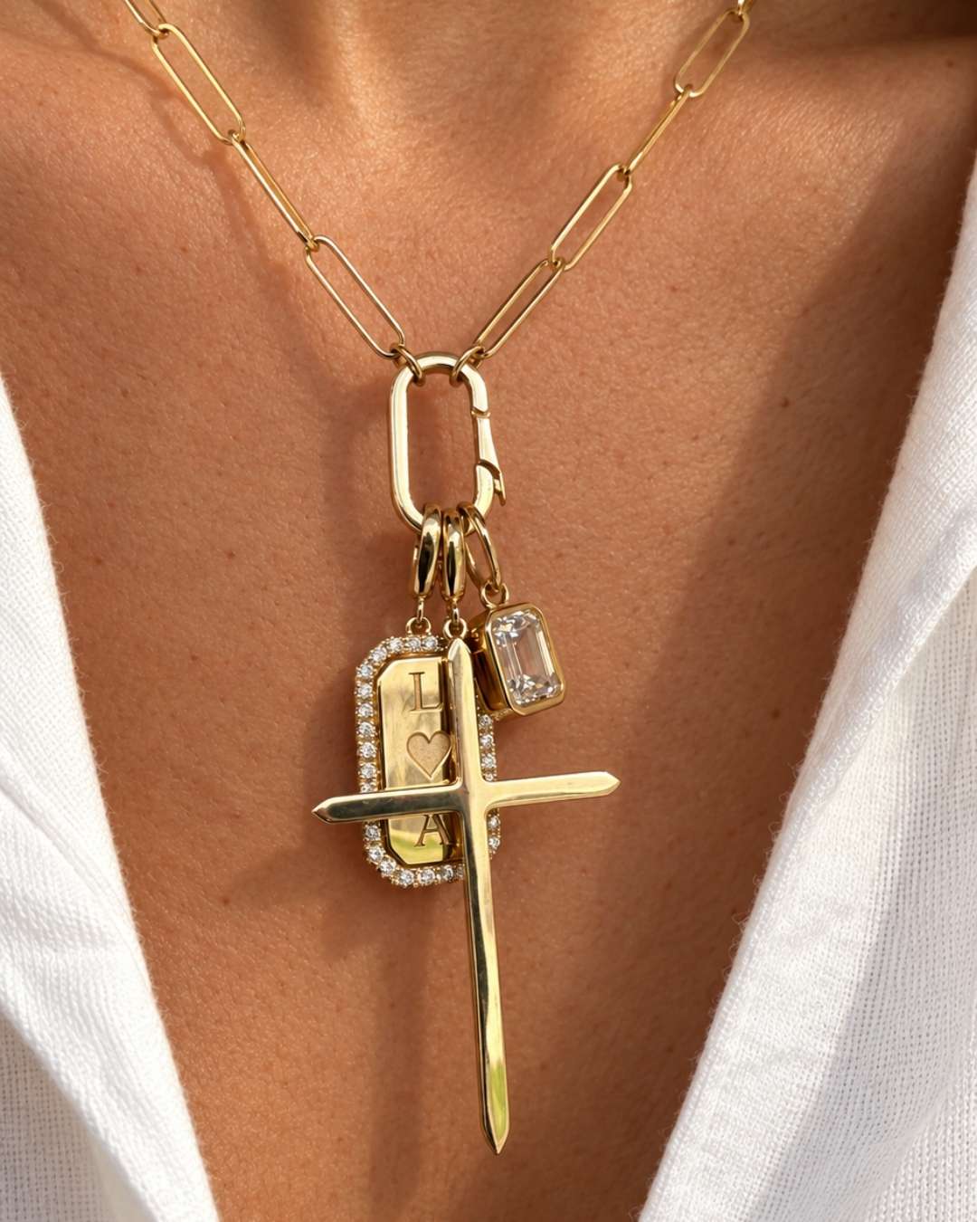 A close-up of a 14K gold necklace worn on a person's neck, featuring a paperclip chain, a rectangular ring clasp, and a multi-pendant charm set with a cross, a emerald cut engraved pendant, and a clear diamond charm.
