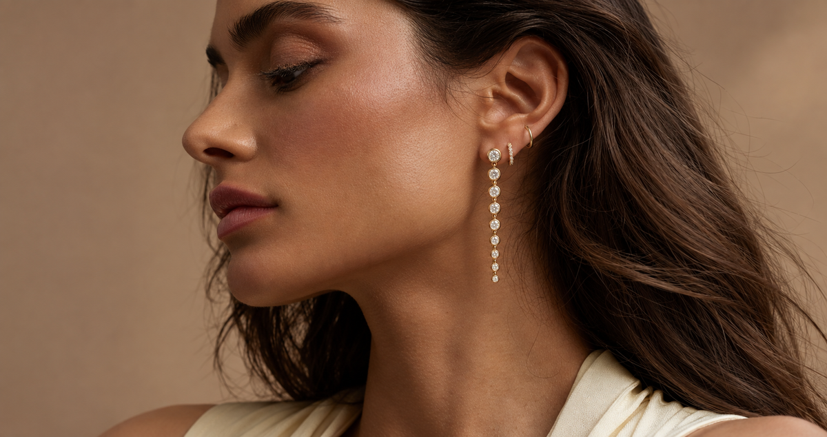 A close-up side profile of a woman wearing a long bezel set diamond graduated dangle earring with small hoops, against a warm beige background for Camille Jewelry.