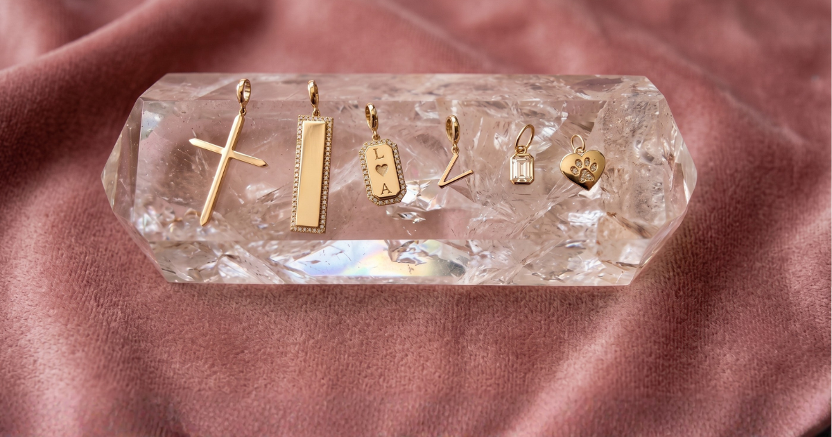 Six gold jewelry for women of solid 14k gold and diamond charms displayed on a clear faceted crystal holder, set against a soft dusty pink fabric background by Camille Jewelry.