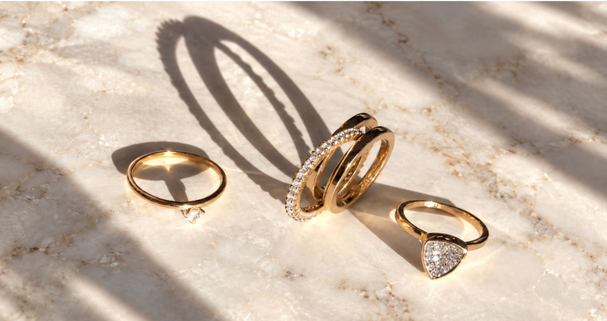 Three gold rings on display. One vermeil ring has a small trillion cubic zirconia setting, one is a double-band insert ring with pavé stones, and one features a trillion concave pavé setting all on a marble display with drastic shadows for Camille Jewelry. 