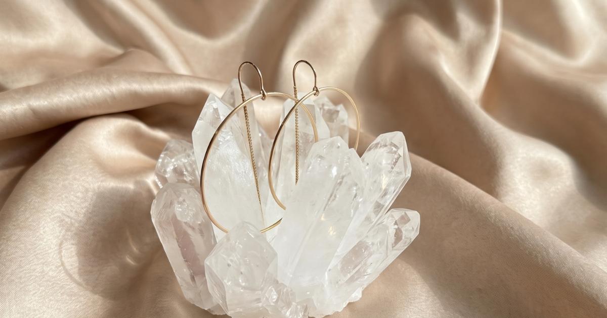 Jewelry gifts under $200 of a pair of threader gold filled hoop earrings displayed on a cluster of clear quartz crystals, set against a draped beige satin fabric background by Camille Jewelry.